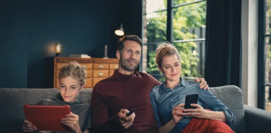 FRITZ!Box: Vodafone bringt Wi-Fi 7 ins Kabelnetz Vater, Mutter und Sohn sitzen auf einem Sofa und nutzen ein Tablet, eine Fernbedienung sowie ein Smartphone