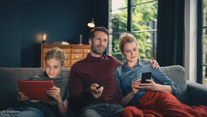 Sommehr_FestnetzAngebot Vater, Mutter und Sohn sitzen auf einem Sofa und nutzen ein Tablet, eine Fernbedienung sowie ein Smartphone